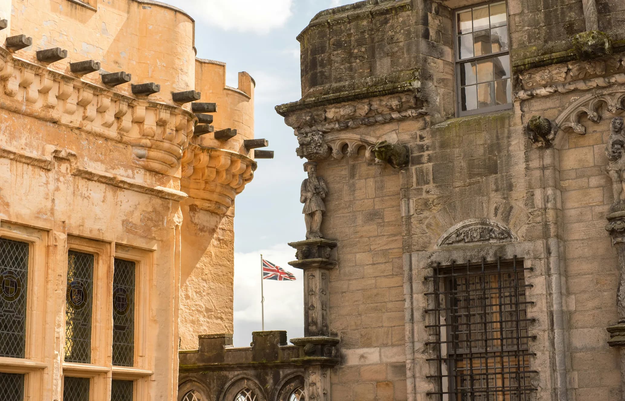 Historic stone castle walls with ornate carvings and windows. A Union Jack flag waves in the background under a partly cloudy sky.