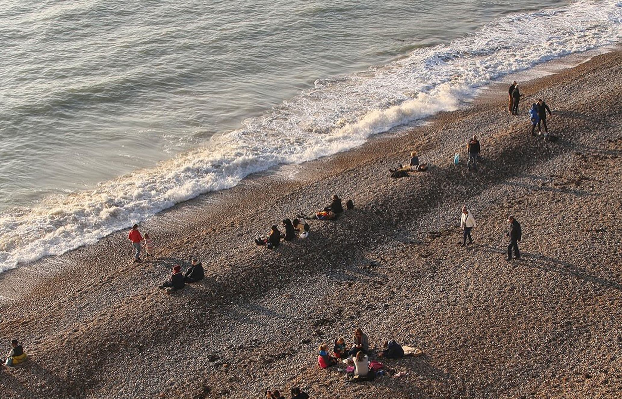 People relaxing and walking on a pebble beach near the shoreline, with gentle waves lapping at the coast.