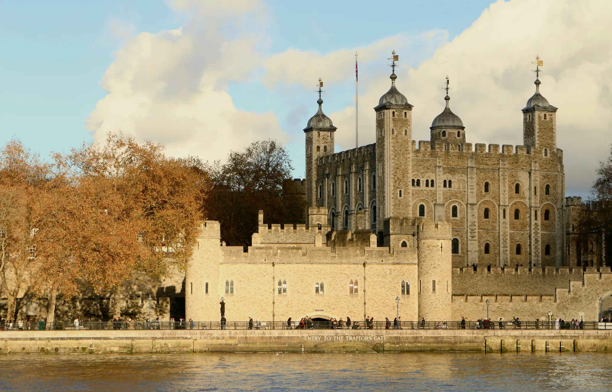 The Tower of London viewed from across the Thames, surrounded by autumn trees, under a partly cloudy sky.