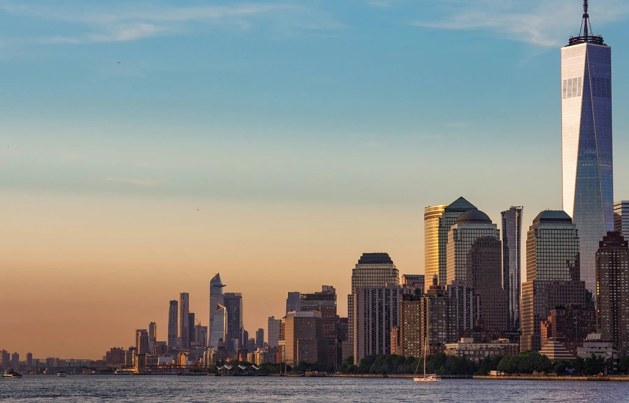 Skyline of a city at sunset with tall skyscrapers and a smooth gradient sky above a calm body of water.