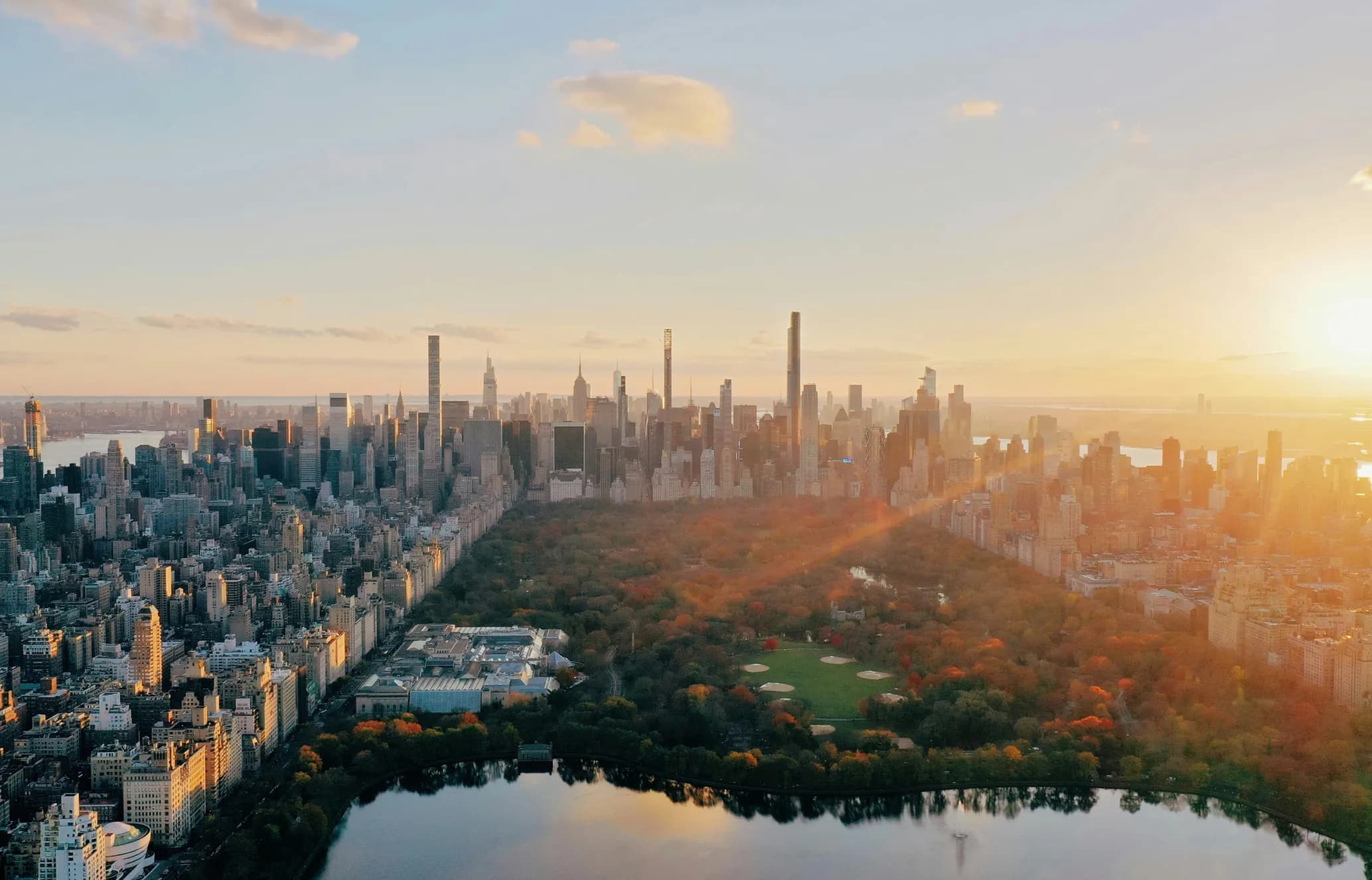 Aerial view of a city skyline at sunset, with a large park and reflecting water body in the foreground.