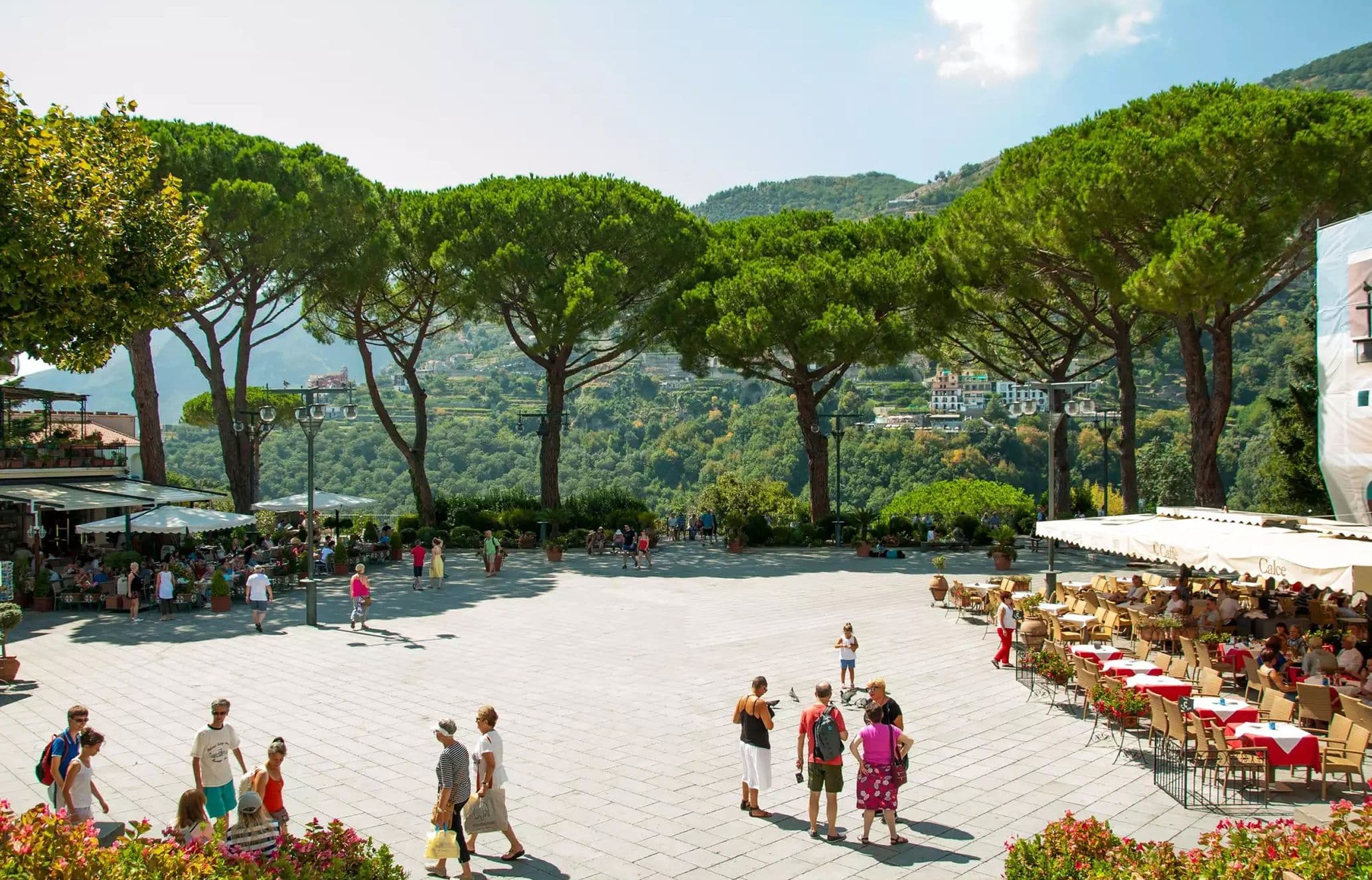 A scenic plaza with people strolling, bordered by umbrella-shaded cafes and framed by tall, lush trees against a mountainous backdrop.