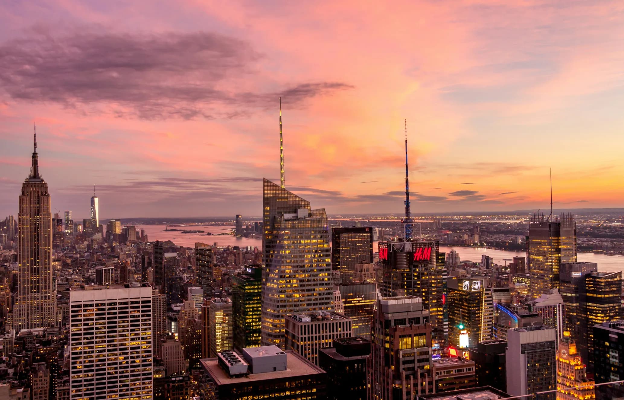 New York City skyline at sunset, showcasing iconic skyscrapers under a vibrant pink and orange sky, with water in the background.