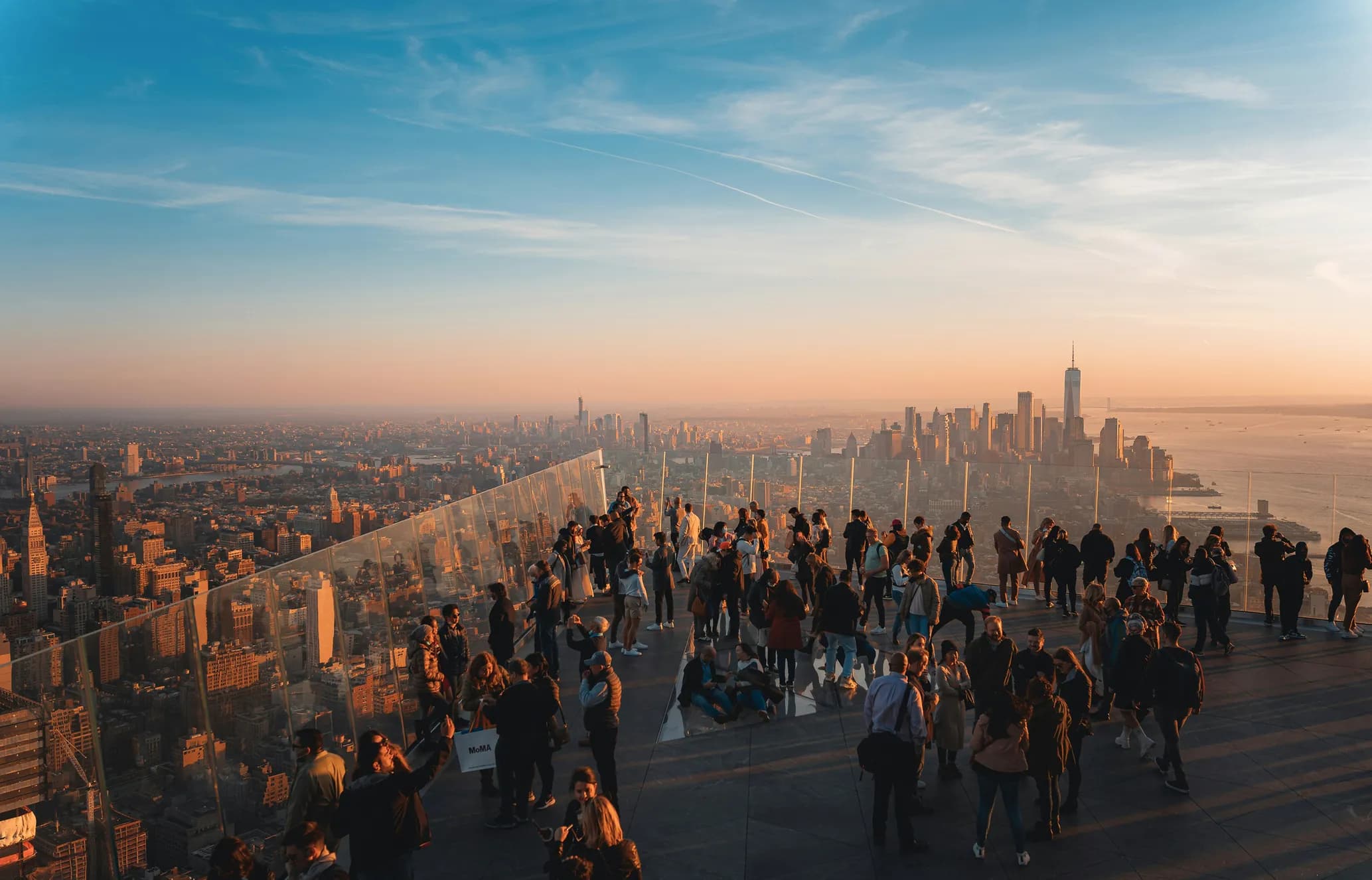 People gather on a rooftop observation deck overlooking a cityscape at sunset, with a vibrant sky and skyscrapers visible in the distance.