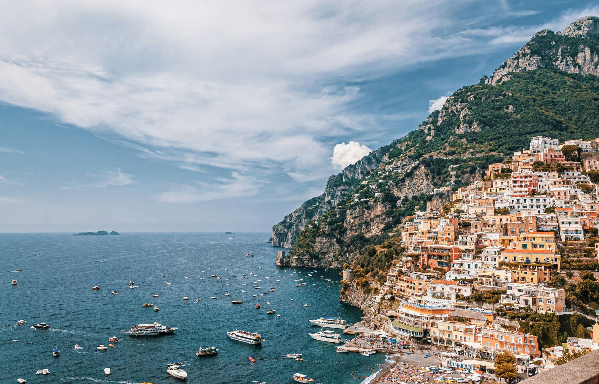 Scenic view of Positano, Italy with colorful hillside buildings, a blue sea dotted with boats, and mountains under a partly cloudy sky.