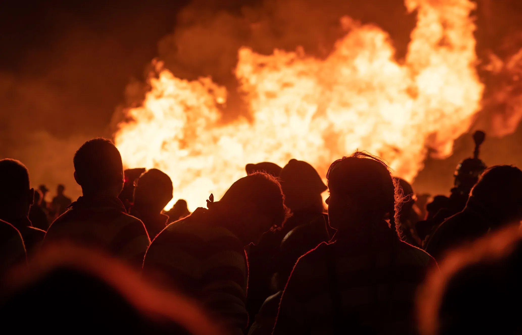 Silhouettes of people gathered around a large bonfire at night, with bright flames illuminating the scene.