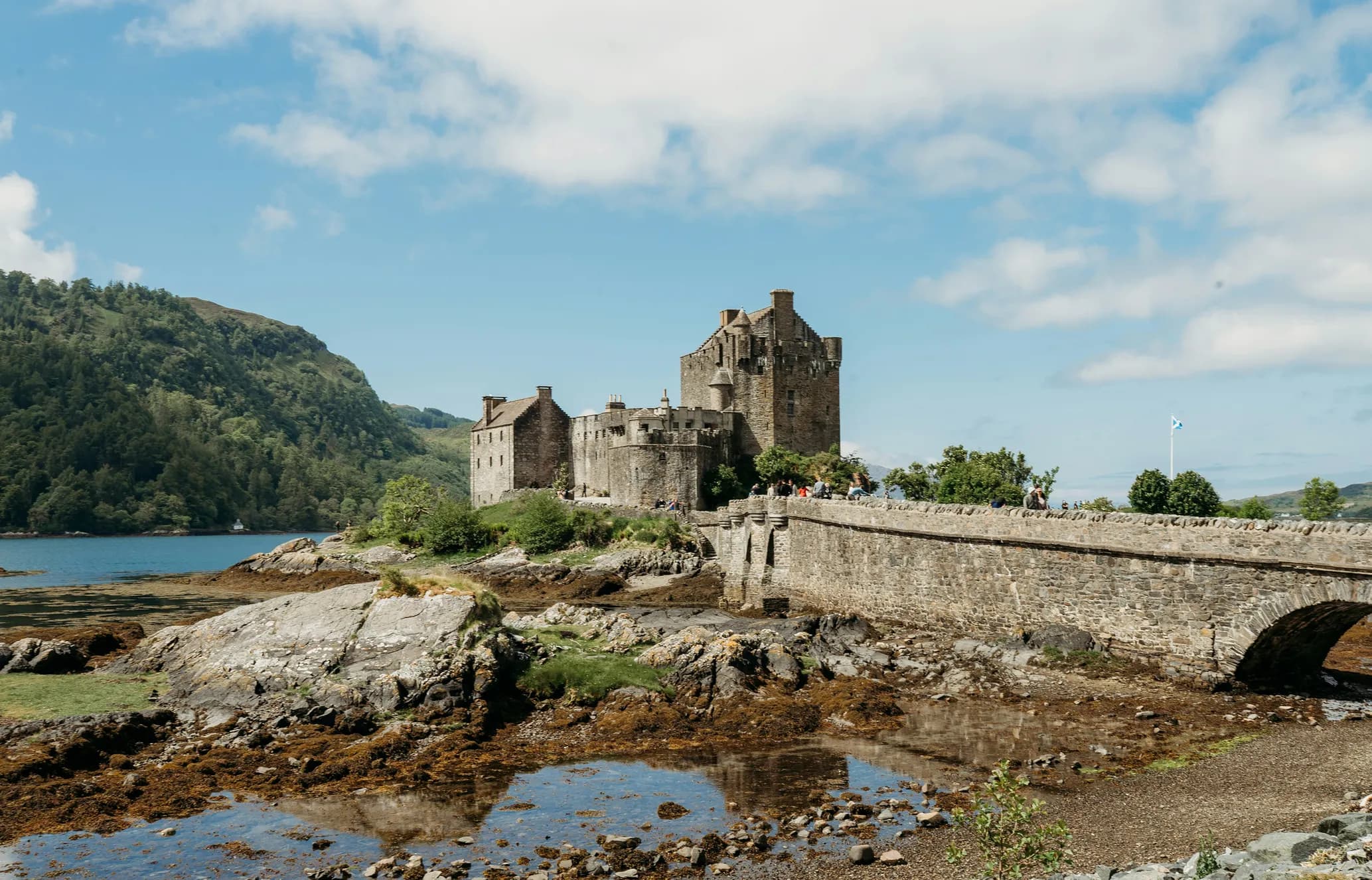 Stone castle with crenelations sits on a rocky peninsula, connected by a stone bridge, surrounded by mountains and water under a blue sky.