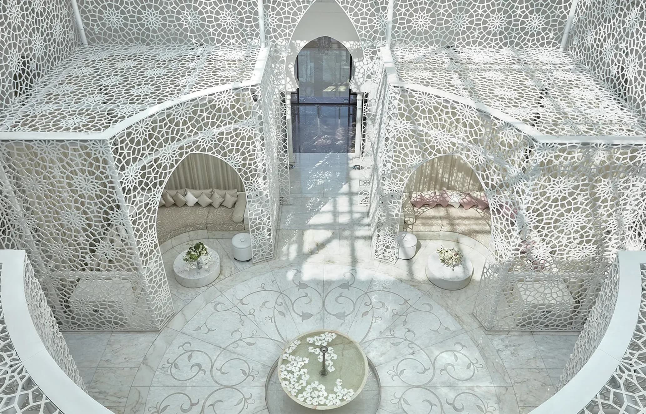 Opulent white interior space with intricate geometric screens, arched seating alcoves, and marble floors patterned with vines. Sunlight casts shadows.