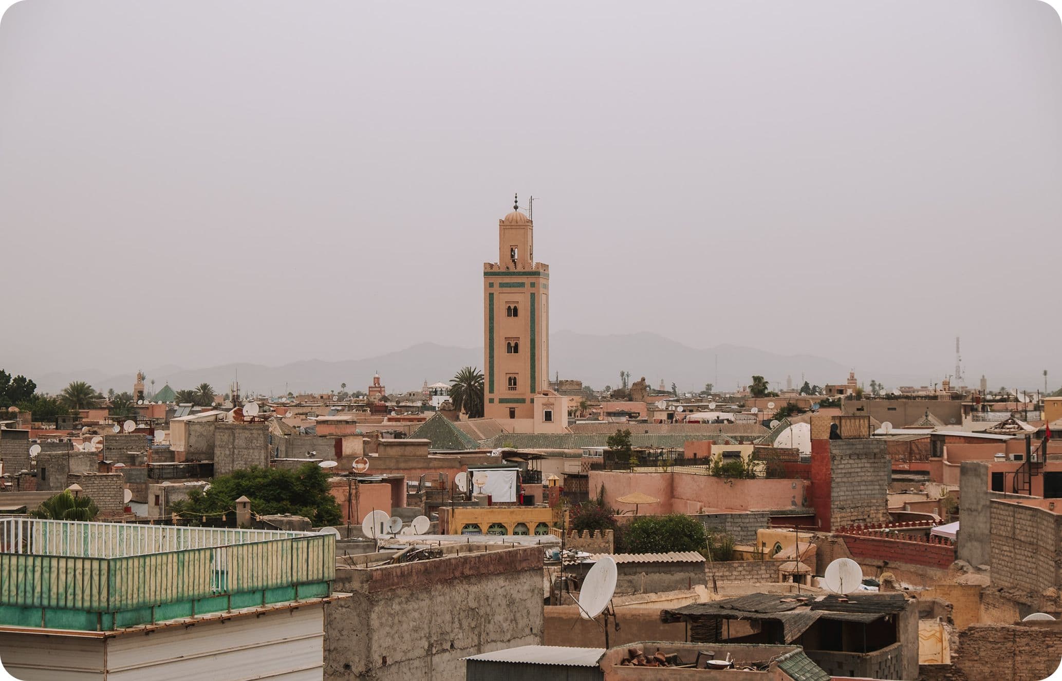 Skyline of a city with a tall, narrow tower in the center, surrounded by low-rise buildings and satellite dishes under a hazy sky.