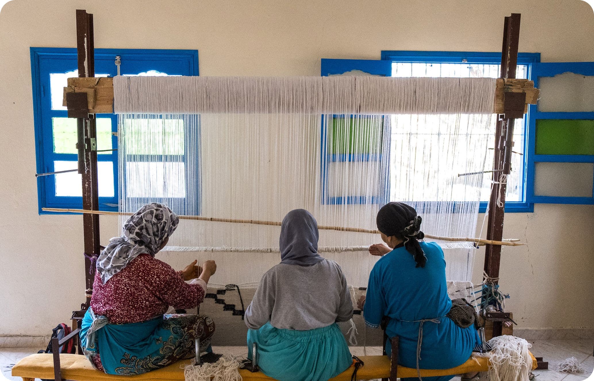 Three women in headscarves and blue garments weave on a large loom in a bright room with blue-framed windows.