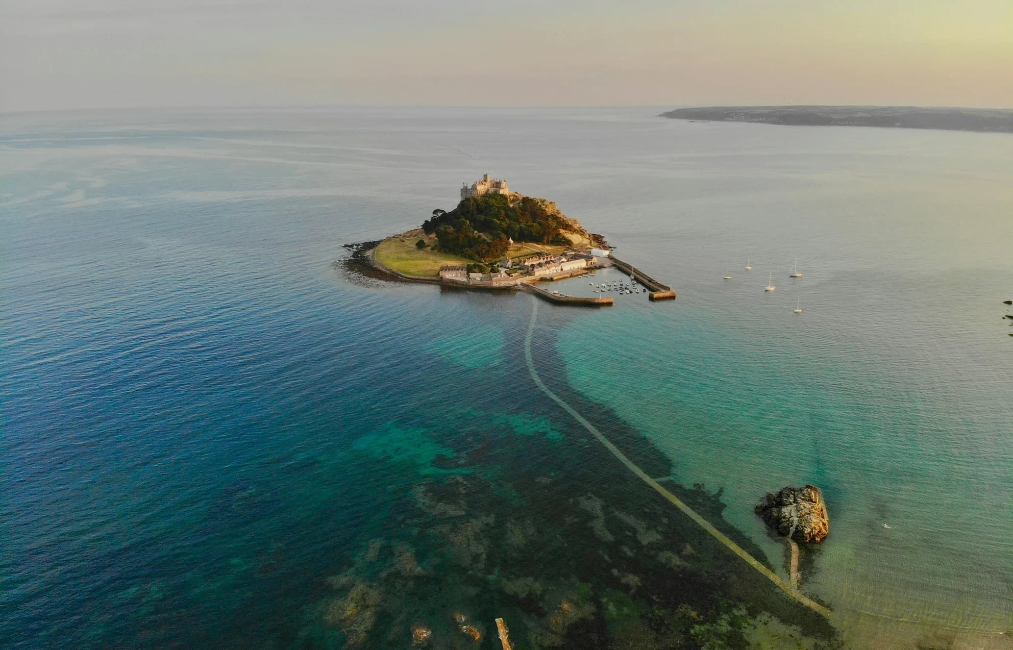 Aerial view of a small, lush island with a castle and a causeway linked to the mainland. Boats are anchored in the surrounding clear blue water.