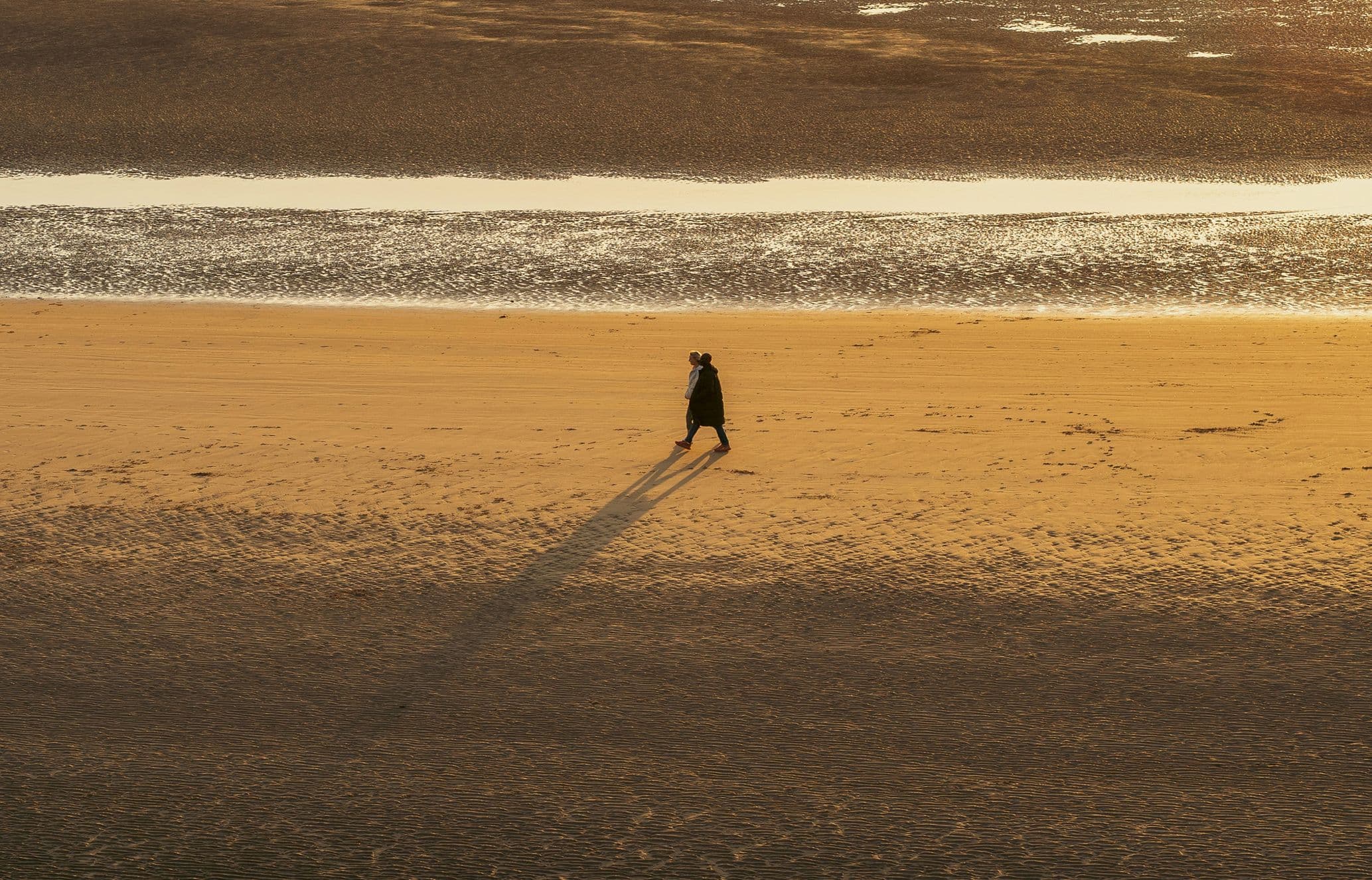 A solitary person walks along a vast, sandy beach casting a long shadow, with a calm sea in the background under soft, warm light.