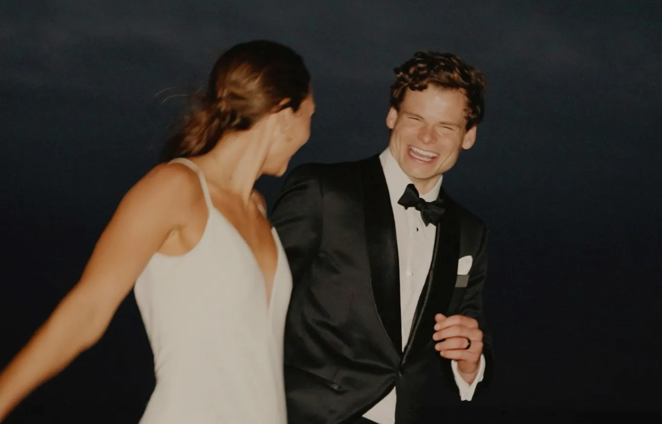 A man in a tuxedo and a woman in a white dress share a joyous moment, smiling and holding hands against a dark background.
