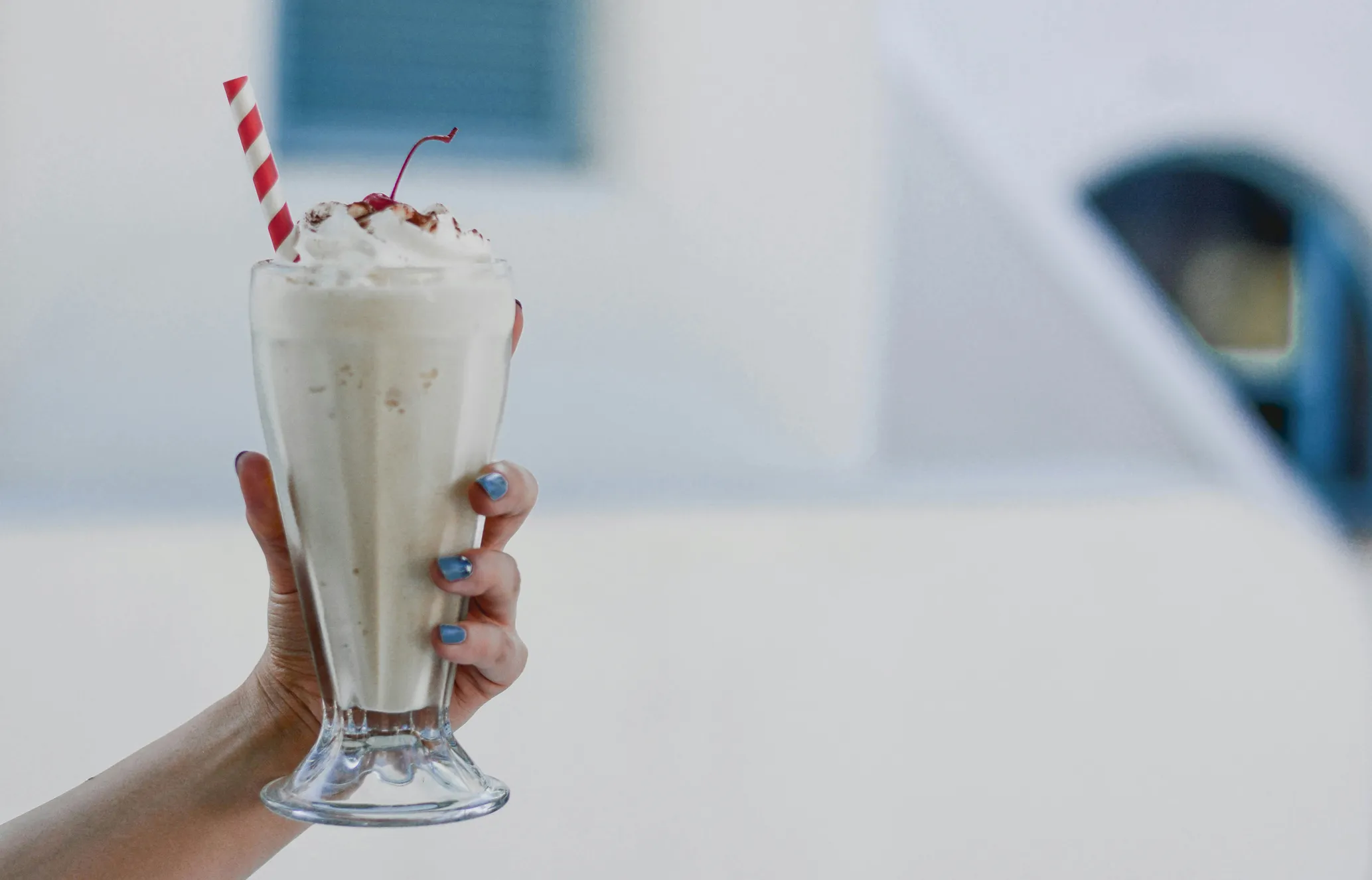 Hand holding a milkshake in a tall glass with whipped cream, a cherry, and a striped straw, with a blurred background.
