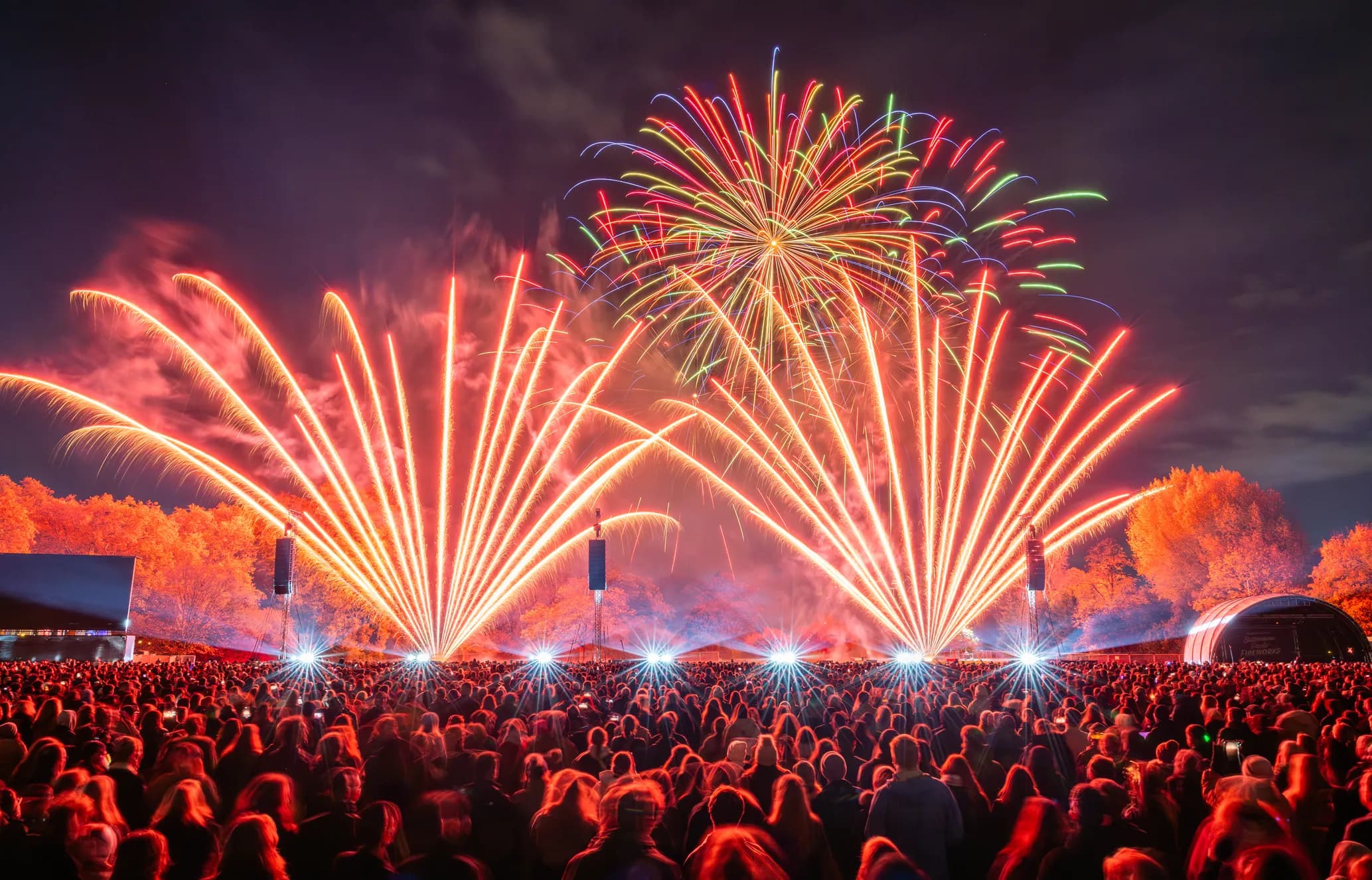A large crowd watches a vibrant fireworks display with red and green bursts against a dark sky, illuminating the scene with bright colors.