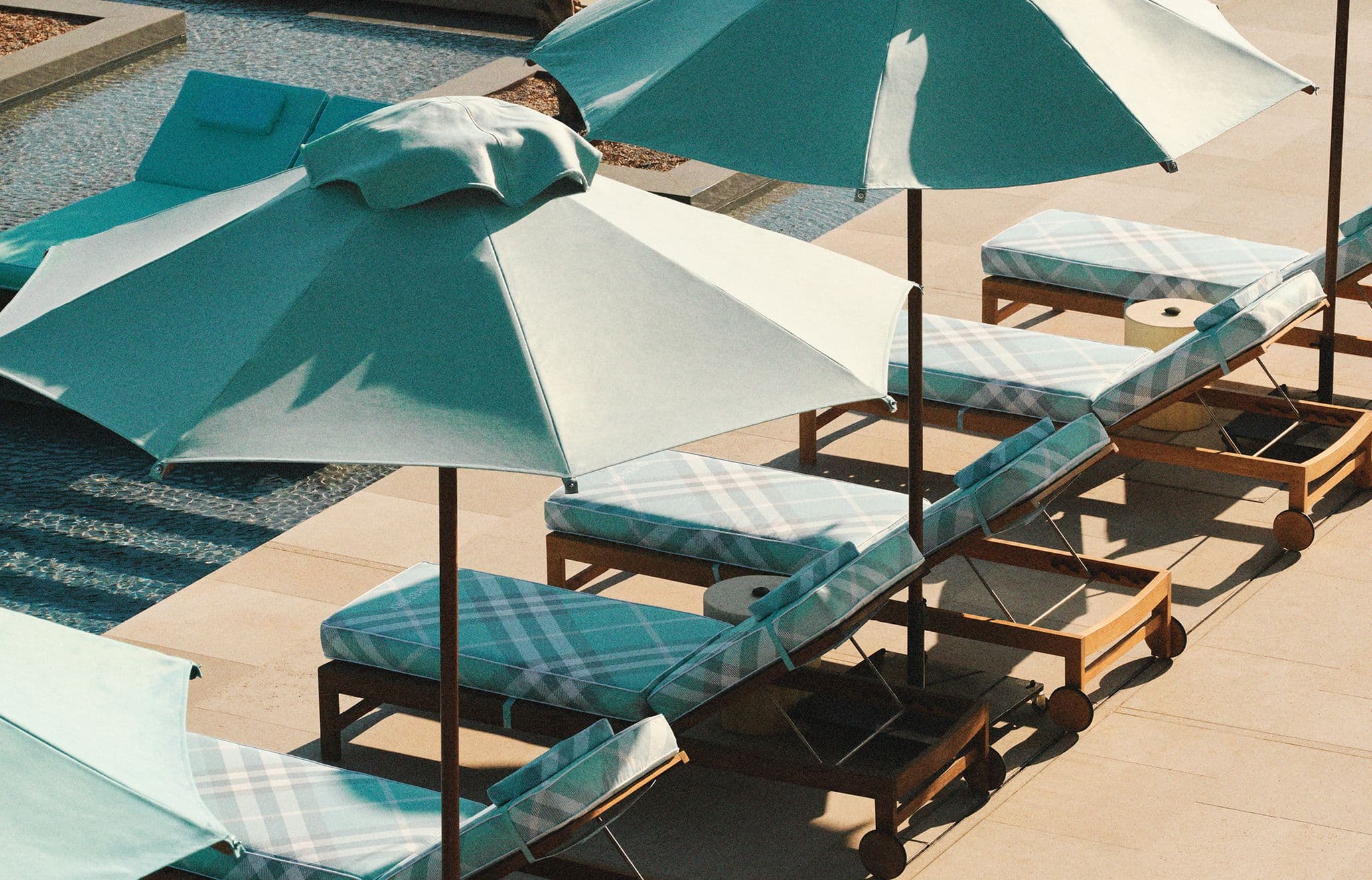 Poolside scene with several lounge chairs covered in plaid fabric, partially shaded by teal umbrellas. Nearby is a swimming pool.