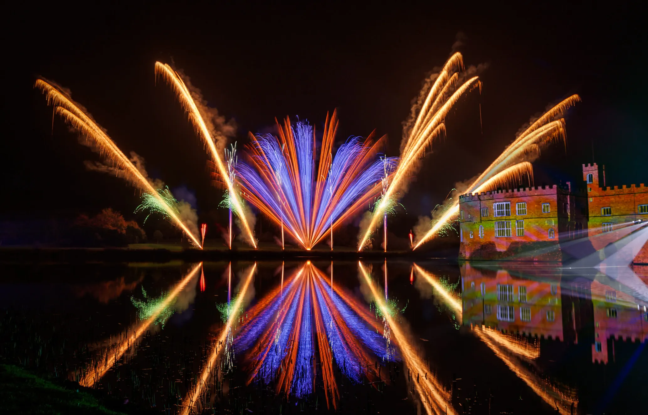 Colorful fireworks burst over a waterway, reflecting brightly, with an illuminated castle in the background against the night sky.