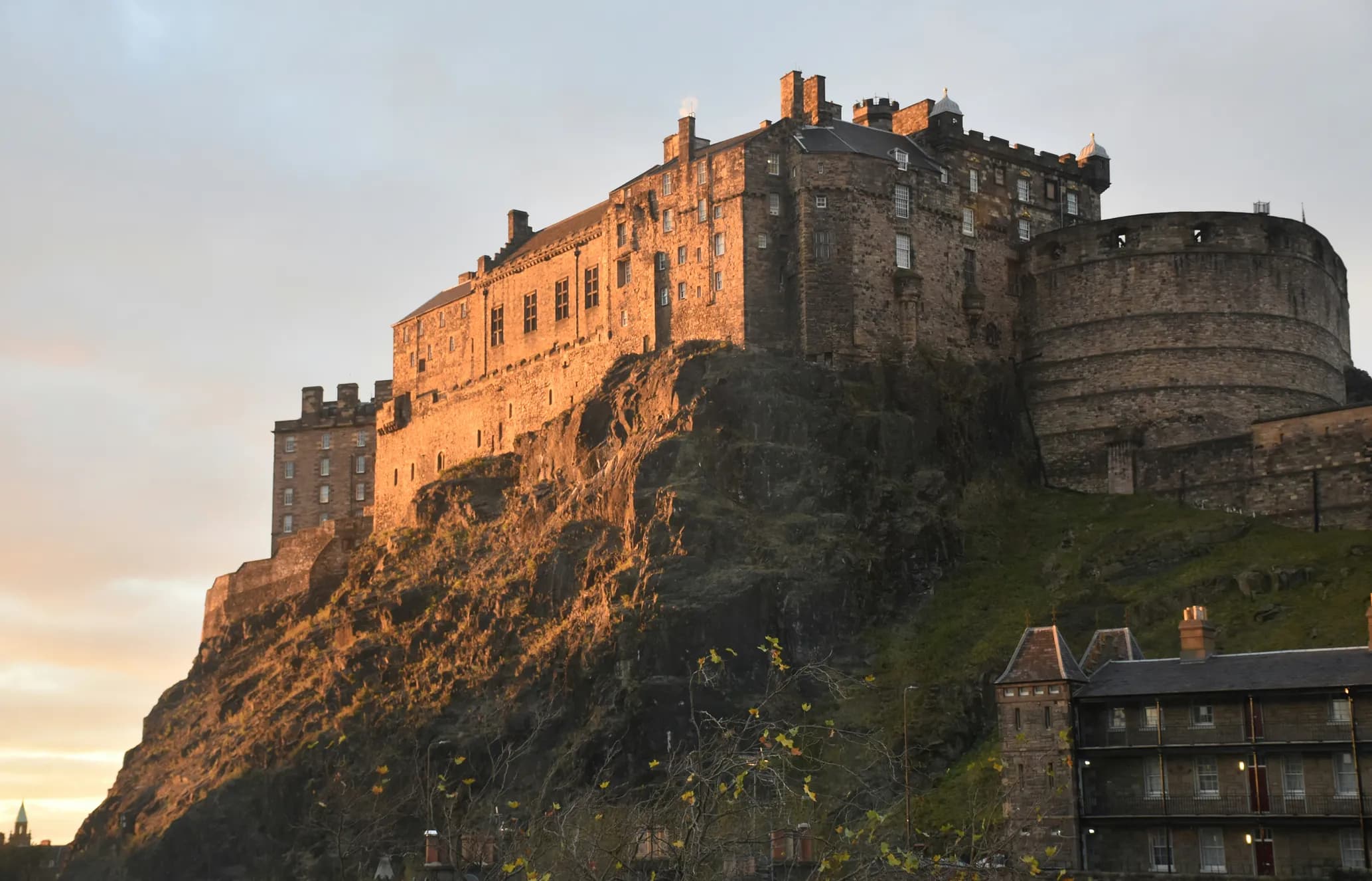 Castle on a rocky hill at sunset, with warm sunlight illuminating the stone walls and a cloudy sky in the background.