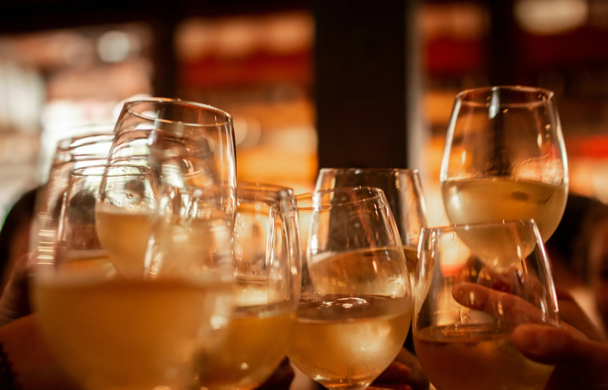 People clinking glasses of white wine in a celebratory toast, with a warm, blurred background suggesting an indoor gathering or event.
