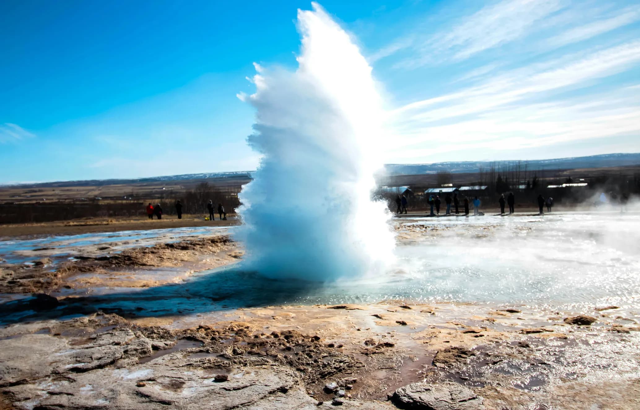 A powerful geyser erupts a tall plume of white water into a bright blue sky, with spectators watching from the wet, rocky terrain.
