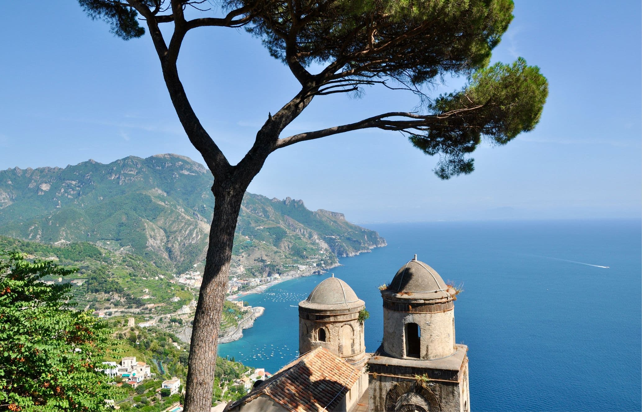 Scenic view of a coastal landscape with a tree and historic buildings overlooking the blue sea and mountains in the background.