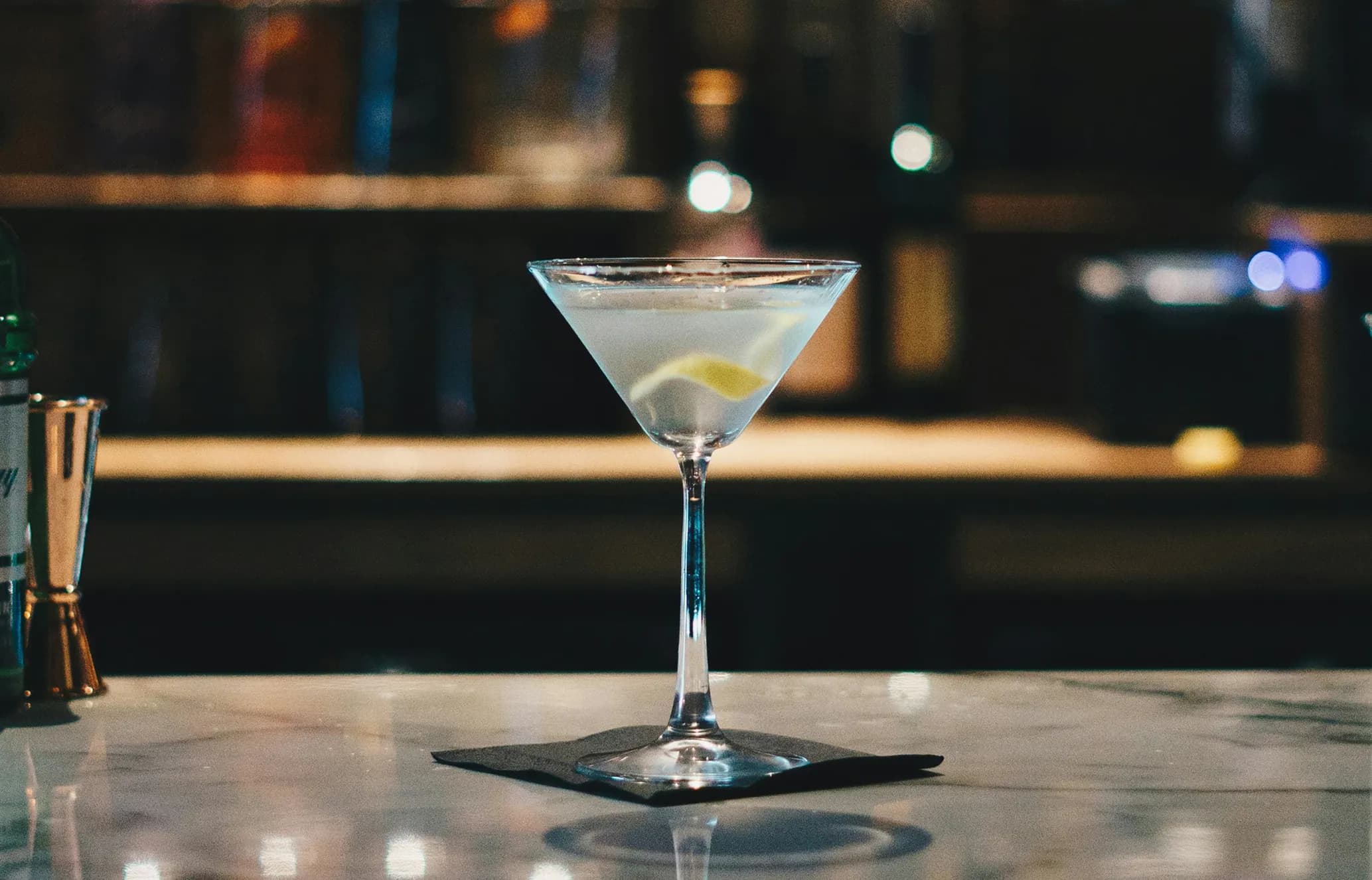 A martini glass filled with a clear cocktail and a lemon twist garnish, placed on a marble bar counter with a blurred background.