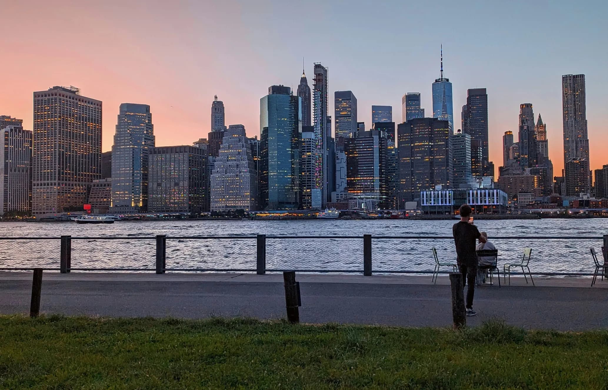 Person taking a photo of the New York City skyline at sunset, viewed from across the water with a clear sky in the background.