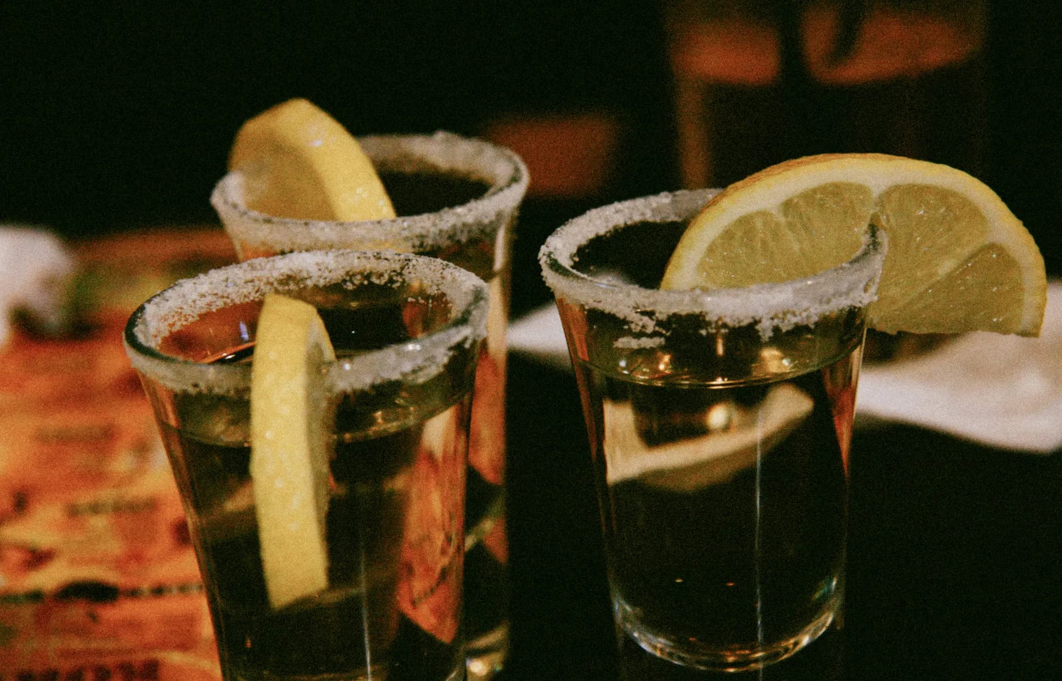 Three shot glasses with salted rims, each topped with a lemon wedge, are placed on a dark surface.