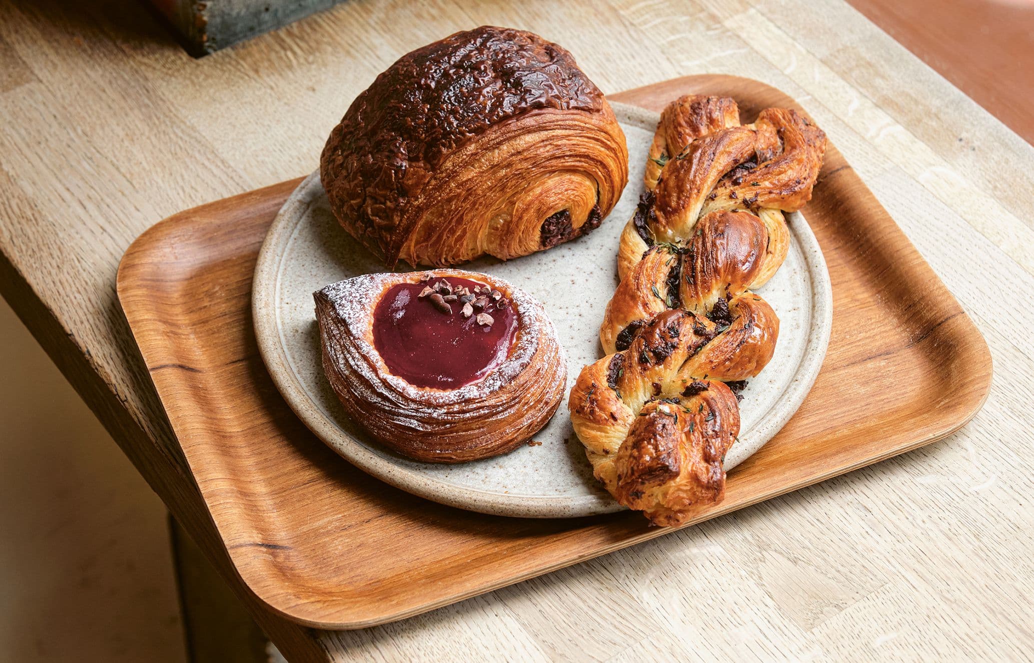 Assorted pastries on a wooden tray: a chocolate croissant, a berry tart with powdered sugar, and a braided pastry with chocolate chips.