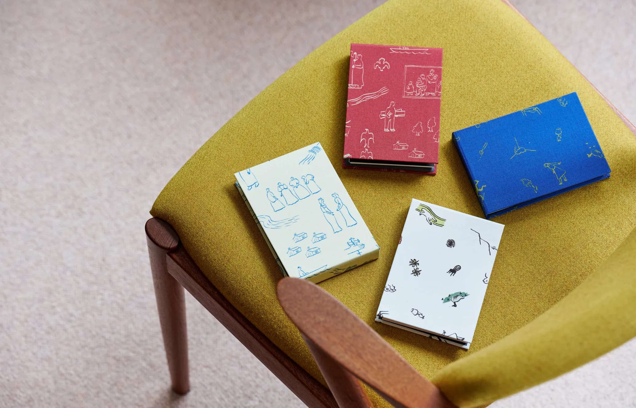 Four small illustrated books in red, blue, white, and light green, featuring various figures and scenes, resting on a mustard yellow chair.