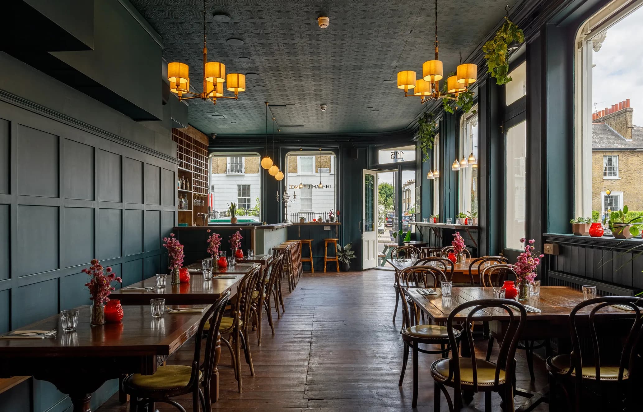 Cozy restaurant interior with wooden tables, chairs, decorative red vases with flowers, warm lighting, and large windows overlooking a street.