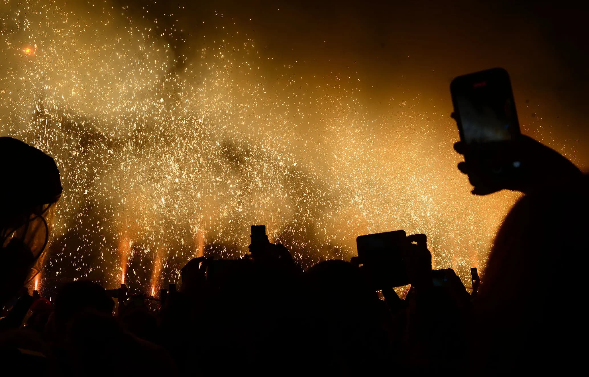 A crowd silhouetted against bright fireworks, with several people holding up phones to capture the display.