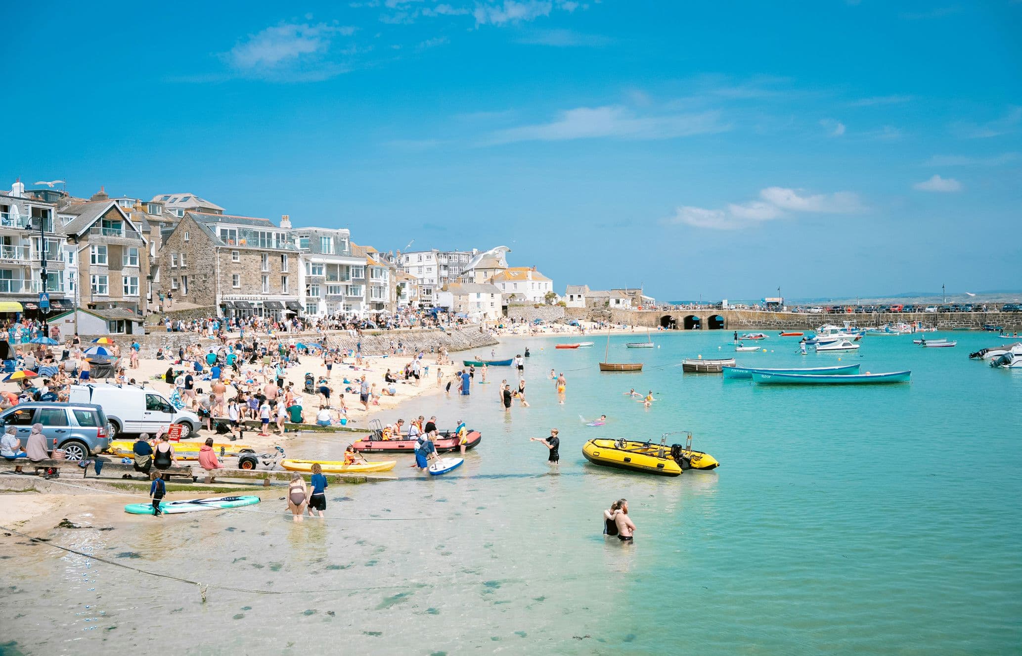Busy beach scene with people sunbathing and swimming near boats in clear turquoise water, with scenic seaside buildings in the background.