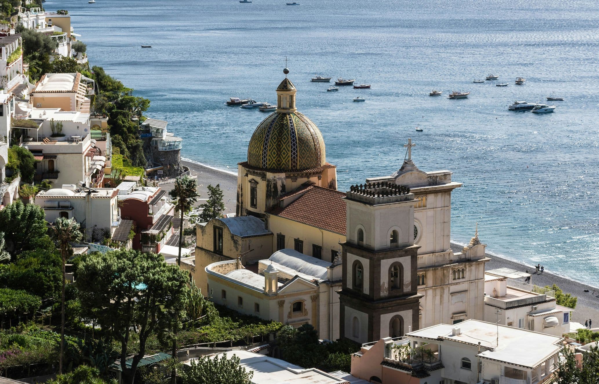 Coastal village with a domed church, colorful buildings, and boats on the sea, nestled along a lush hillside.