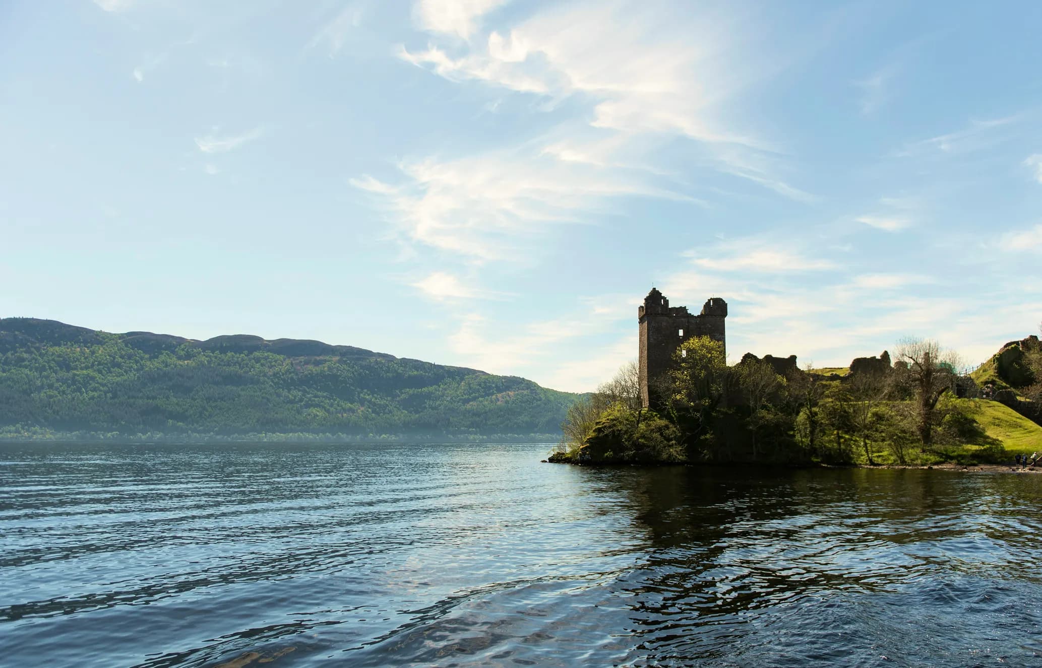 Ruins of a castle tower on a hill beside a tranquil lake, with forested mountains and a partly cloudy blue sky in the background.