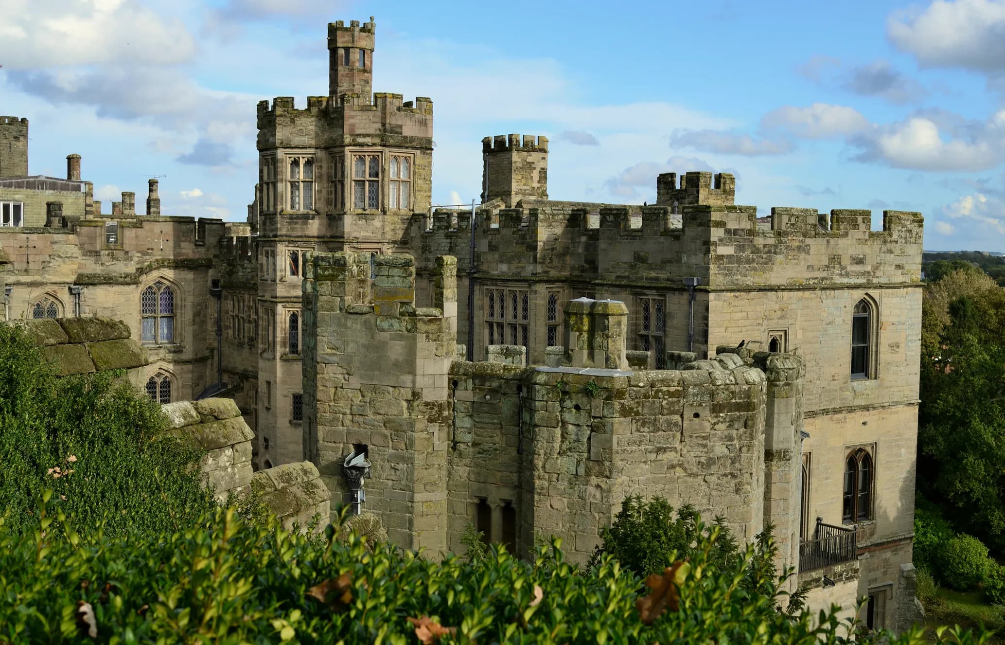 A historic stone castle with crenelated towers and arched windows set against a partly cloudy blue sky, surrounded by greenery.