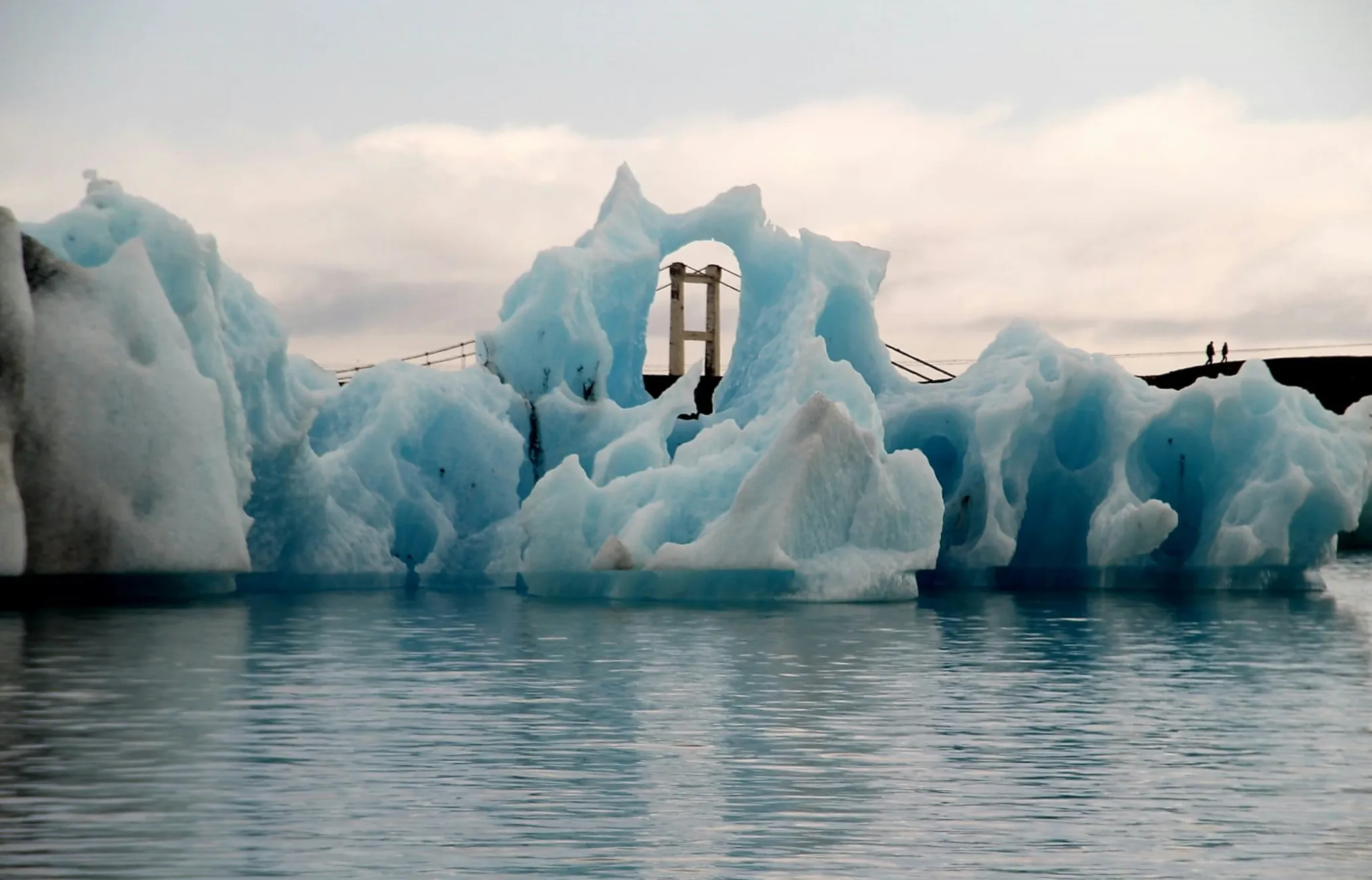 Blue icebergs float in calm water, with one framing a bridge support. Two small figures walk on distant land under a cloudy sky.