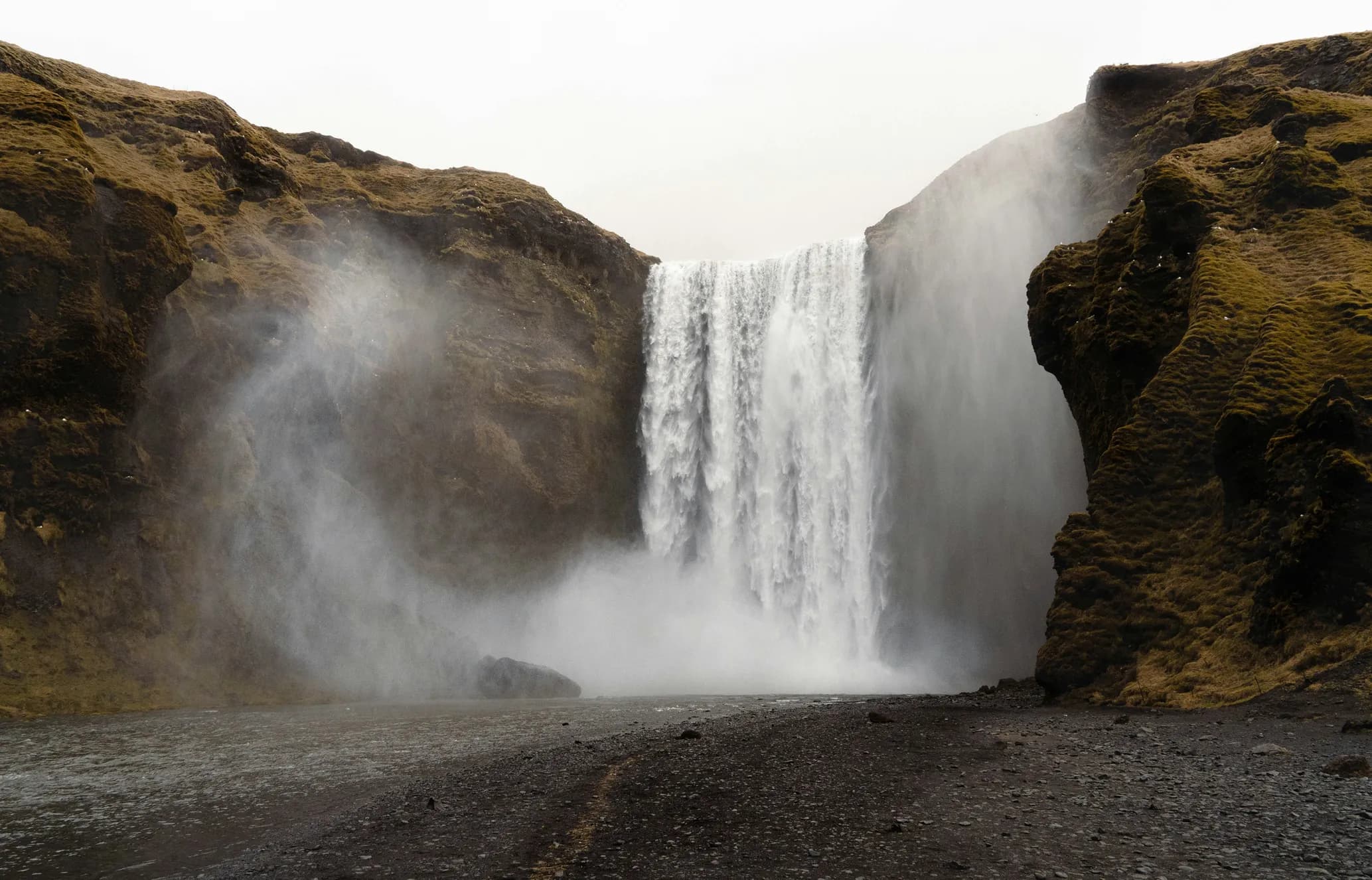 A powerful waterfall cascades between two moss-covered cliffs, with small white birds perched on them. Mist rises from the base over a rocky riverbed.