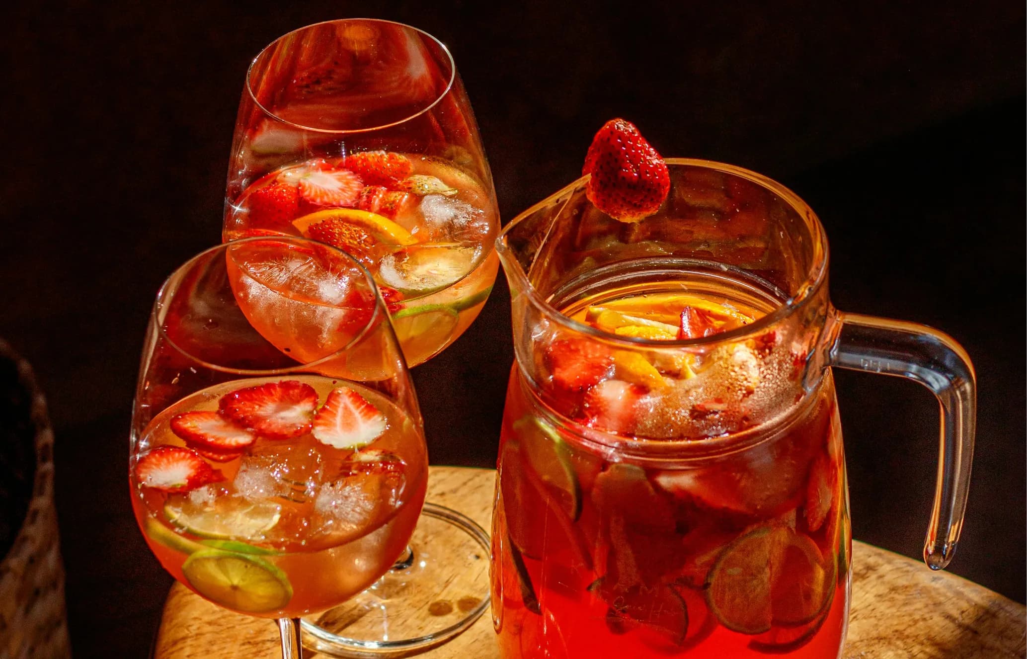 Three glasses and a pitcher of fruity punch with ice, strawberries, and citrus slices in warm lighting on a wooden surface.