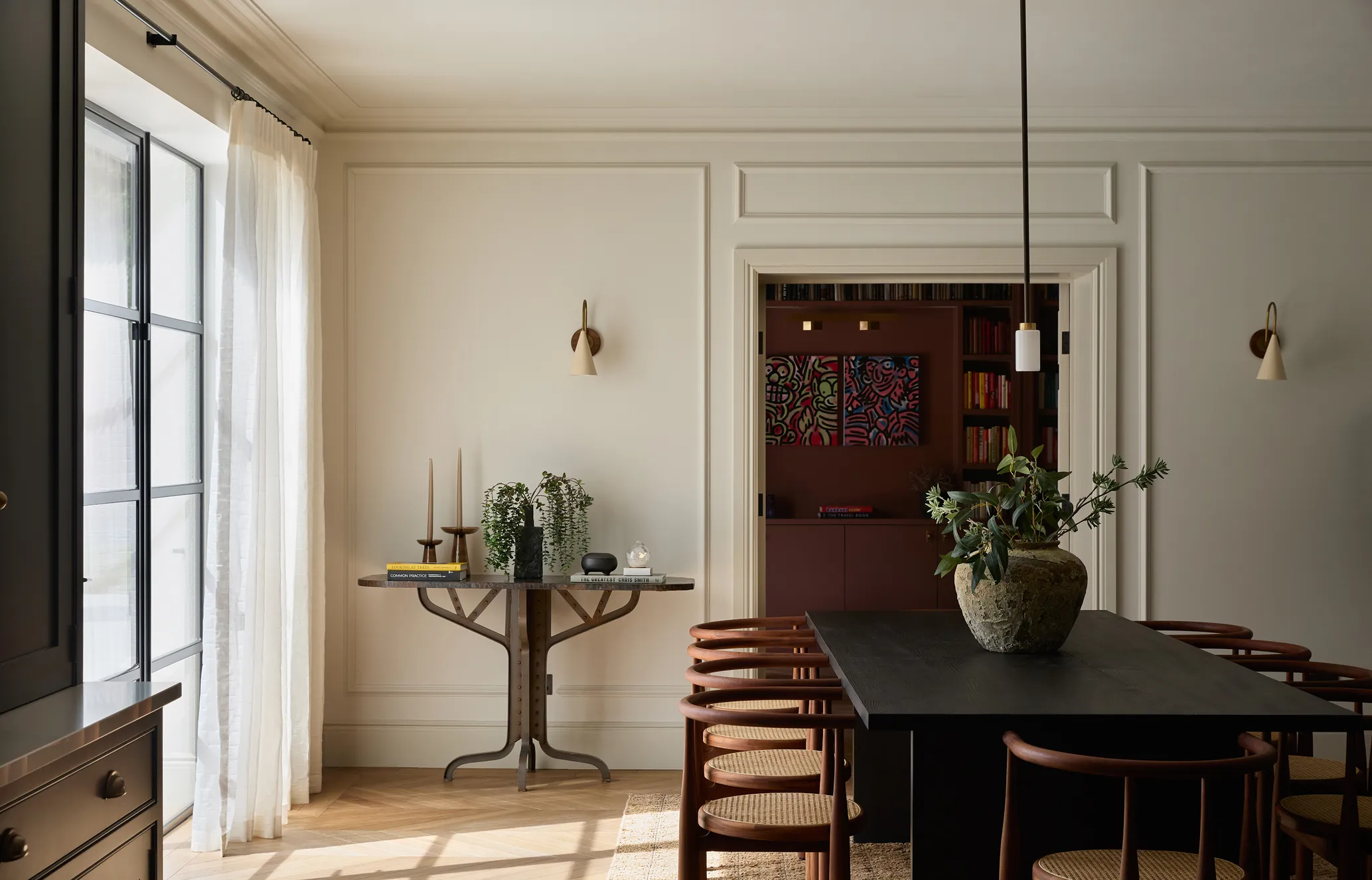 Elegant dining room with black table, wooden chairs, large vase centerpiece, wall sconces, side table, and soft natural light from a window.