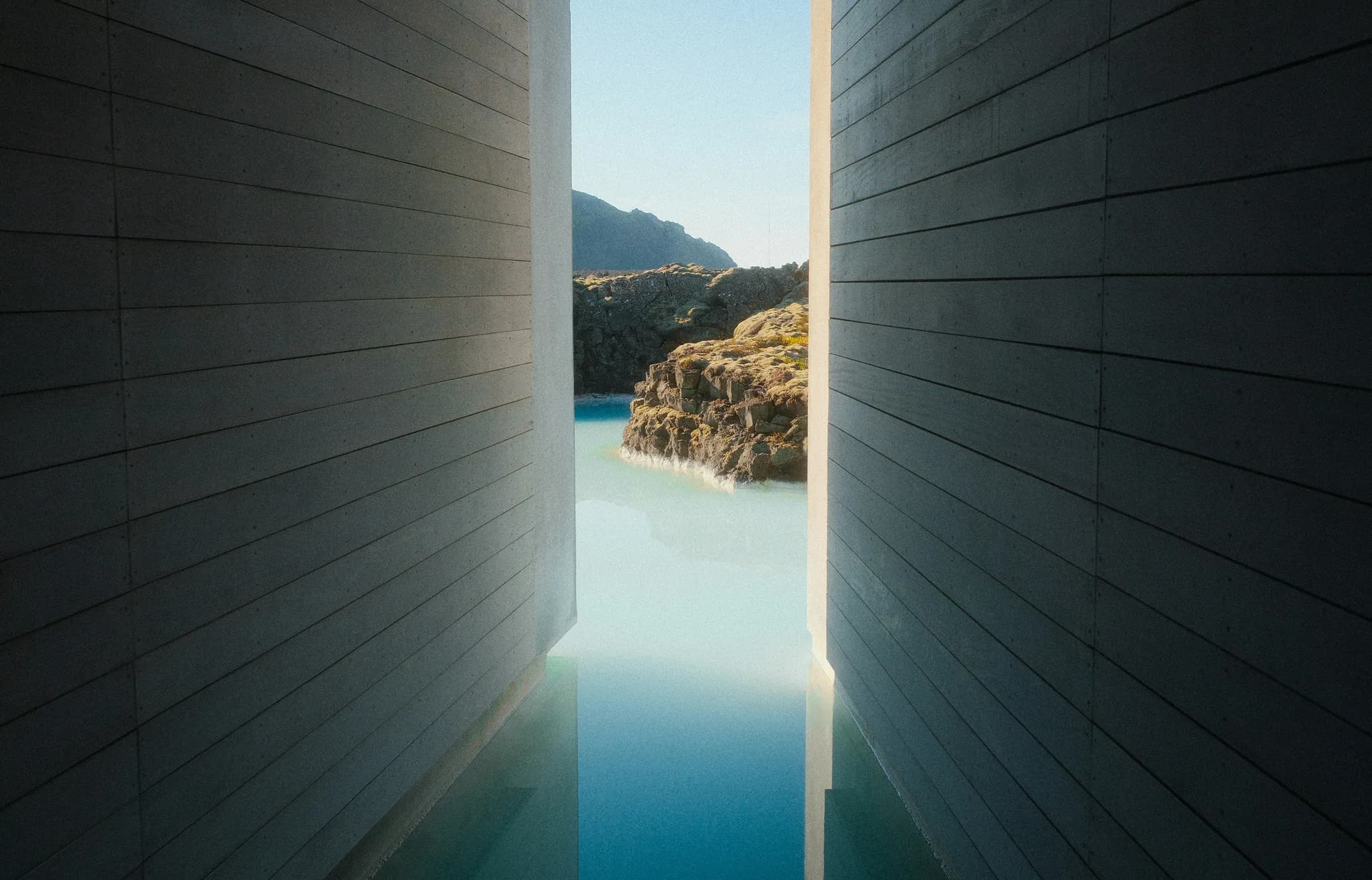 View through a narrow opening between two dark paneled walls, revealing a milky blue geothermal lagoon surrounded by mossy volcanic rocks.