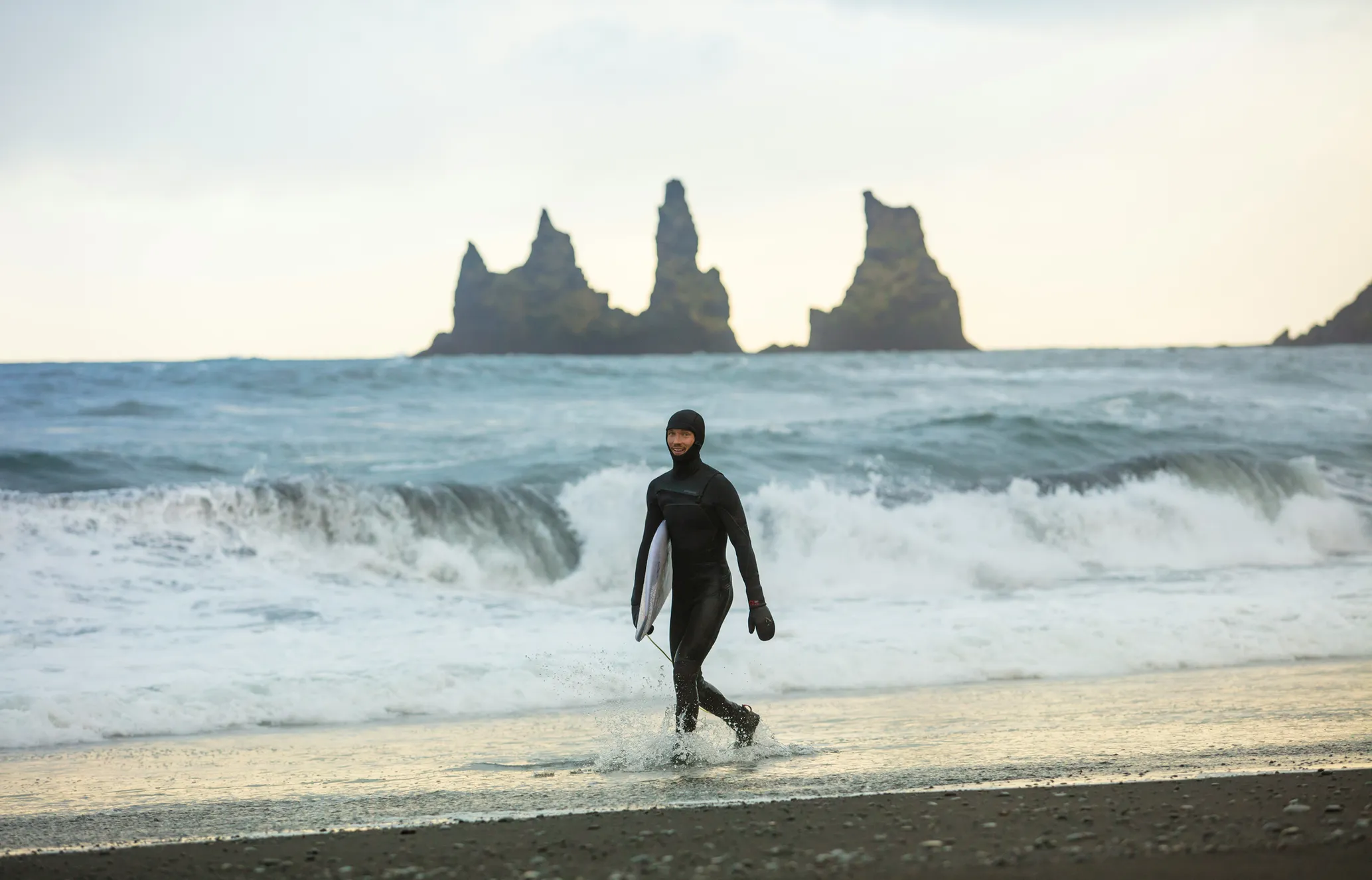A surfer in a black wetsuit walks out of the ocean with his board on a dark sand beach, with large waves and distant sea stacks.