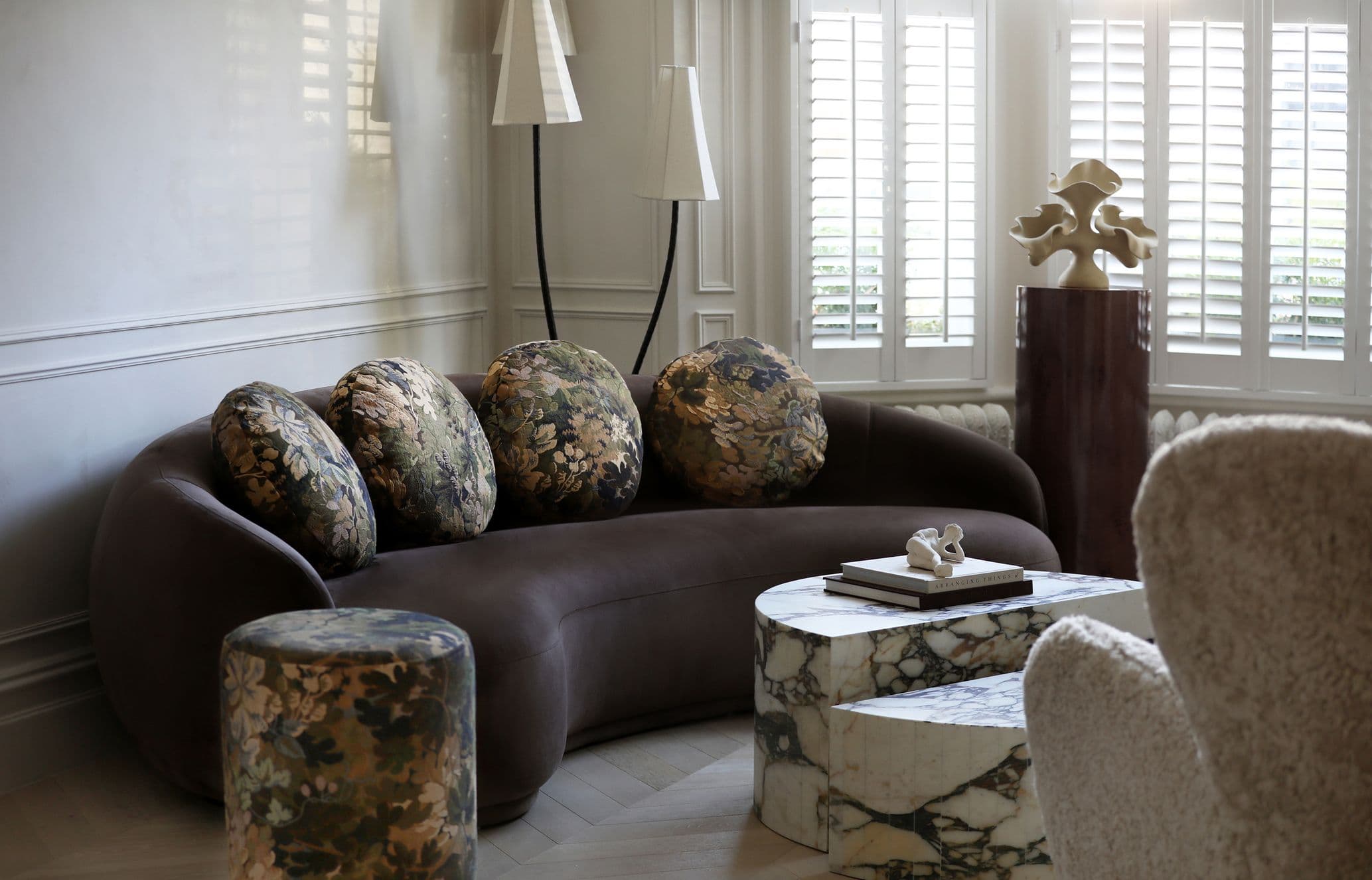 A stylish living room with a brown floral-patterned sofa, marble coffee tables, a sculptural lamp, and a decorative piece on a pedestal.