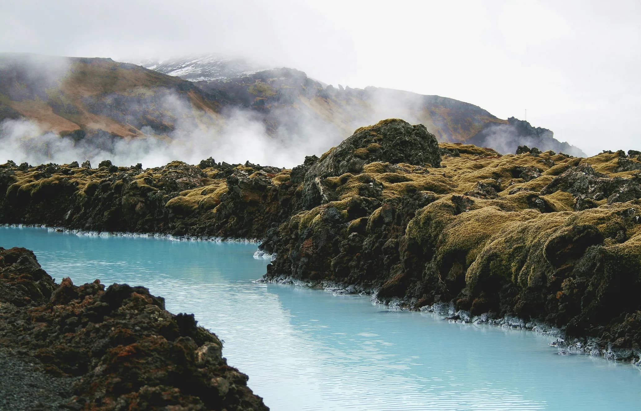 Milky blue geothermal water winds through dark, moss-covered lava fields. Misty mountains and rising steam fill the background.