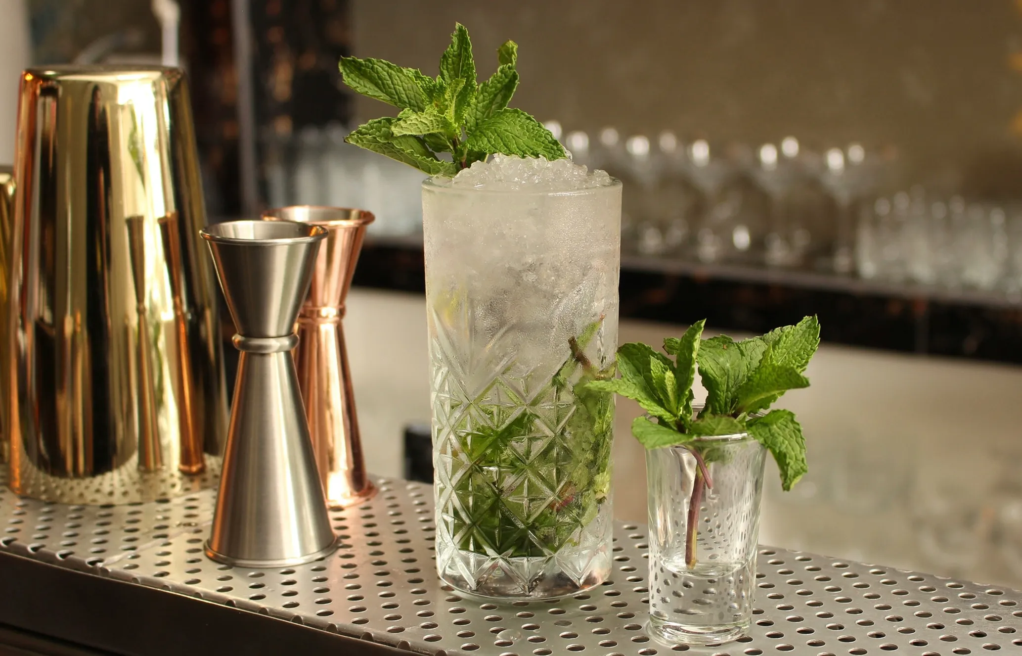 A tall cocktail glass filled with ice and mint leaves, next to a smaller glass with mint, and copper and silver bar tools on a metal counter.