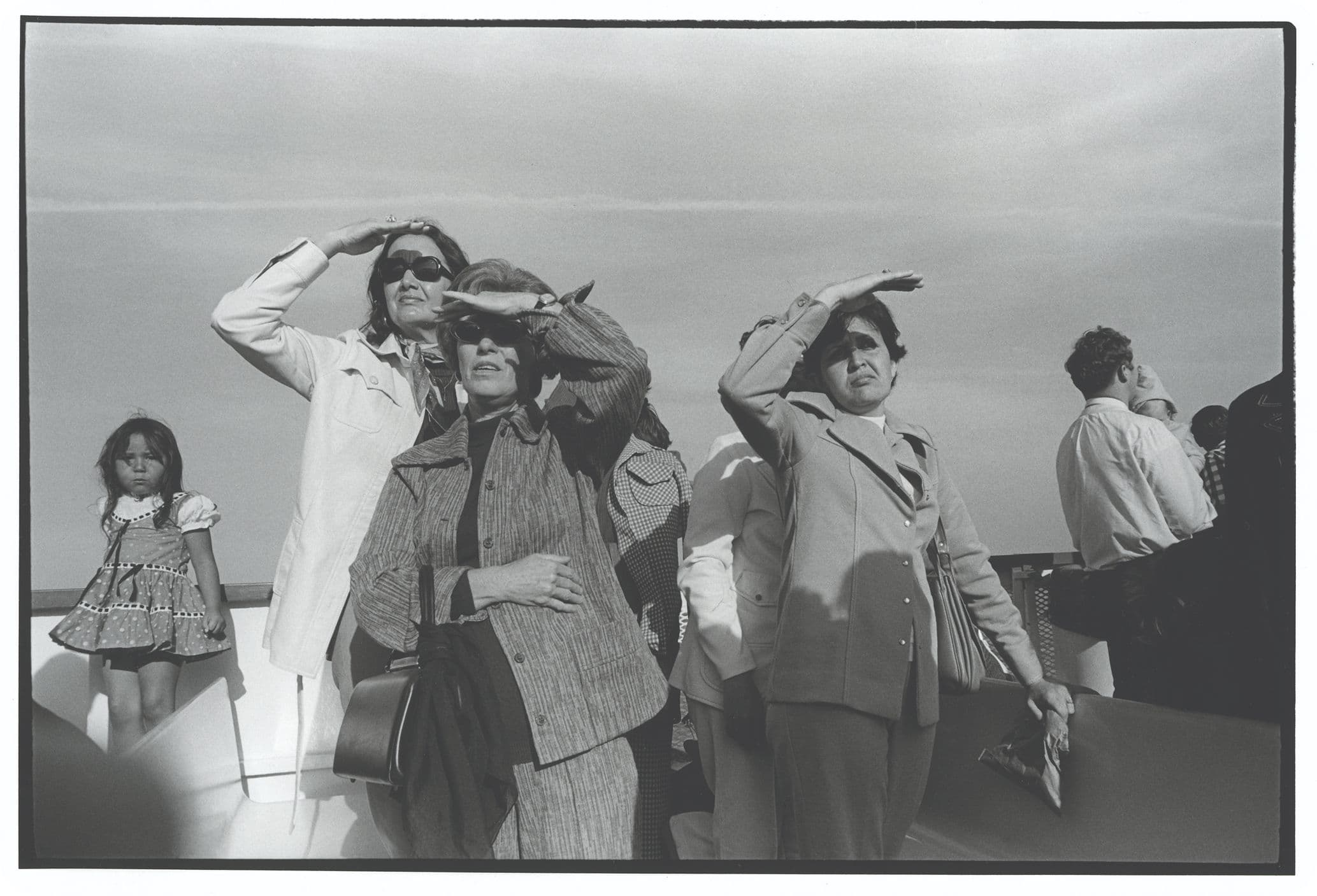 Group of people on a boat, shielding their eyes from the sun, looking into the distance; one child stands nearby. Black and white photo.