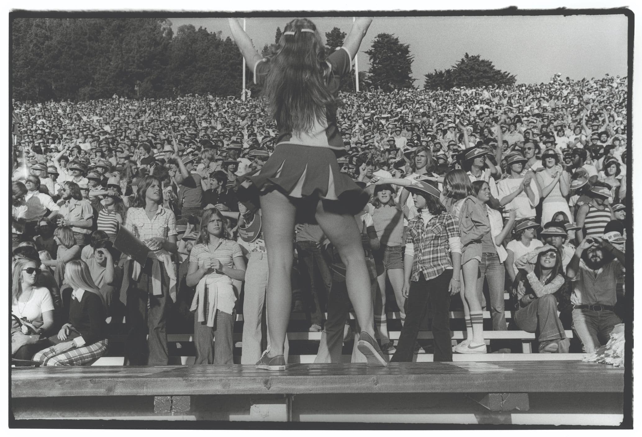 Cheerleader with pom-poms leads a large, cheering crowd in a stadium during a daytime event.