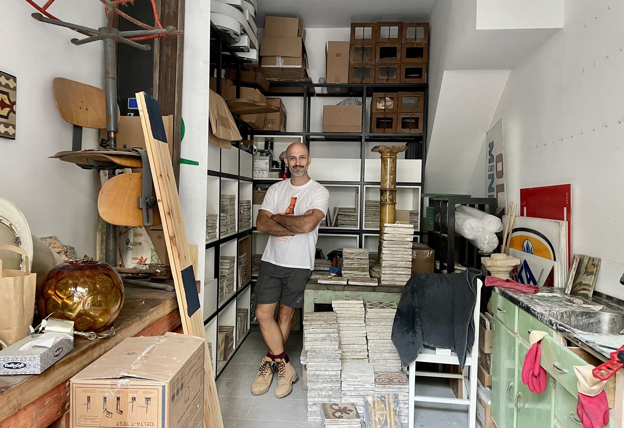 A man stands in a cluttered storage room with shelves of boxes, stacked trays, and various items. He's wearing a T-shirt and shorts, smiling confidently.