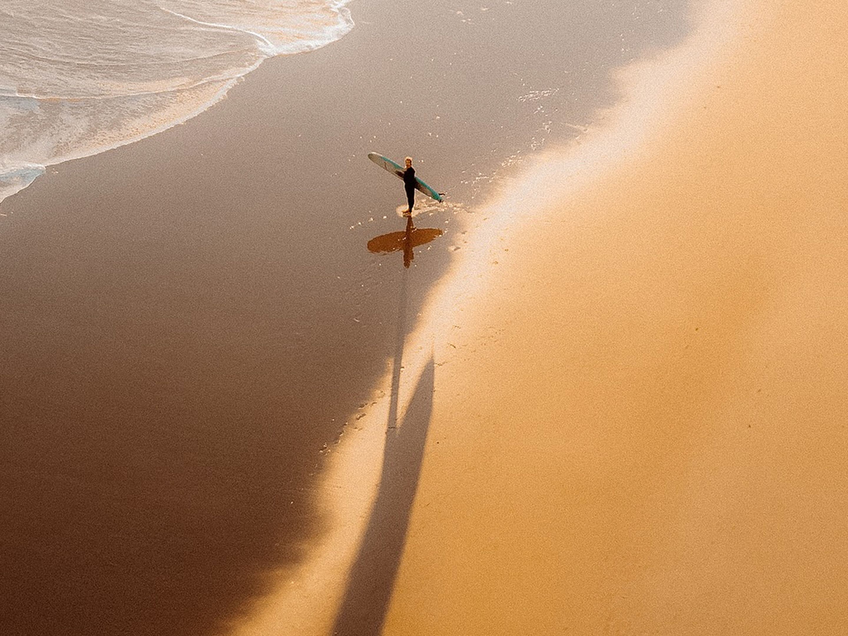 An aerial photogrpah of a surfer approaching the water from a sandy beach, shot from a drone