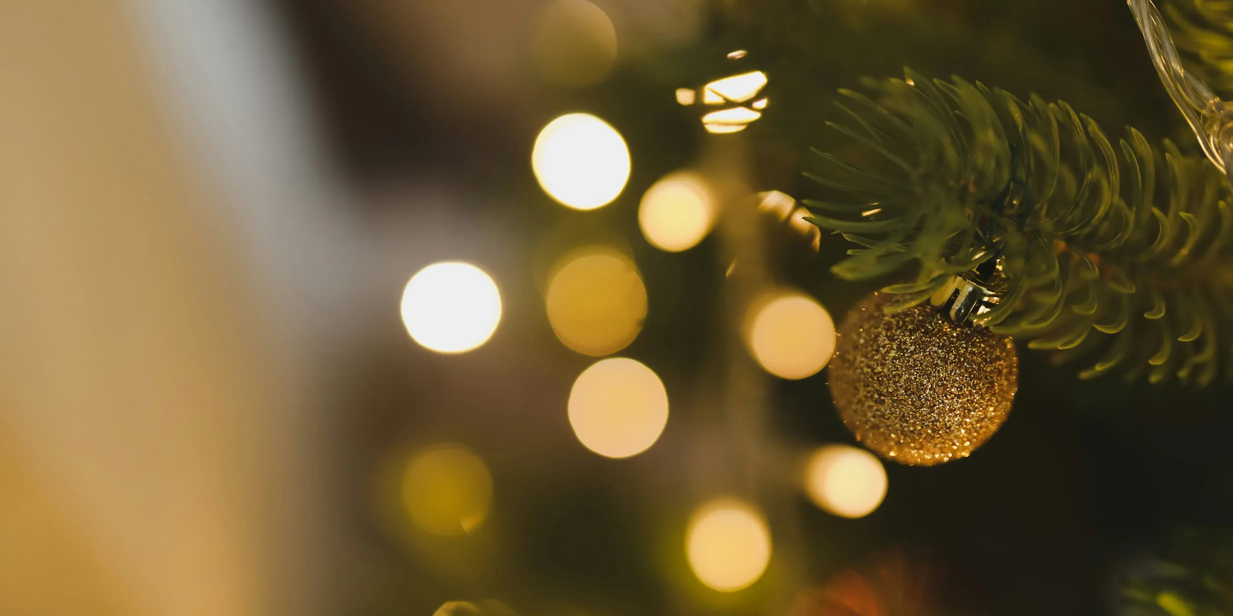 Close-up of a Christmas tree with a gold ornament. Blurred golden lights sparkle softly in the background.